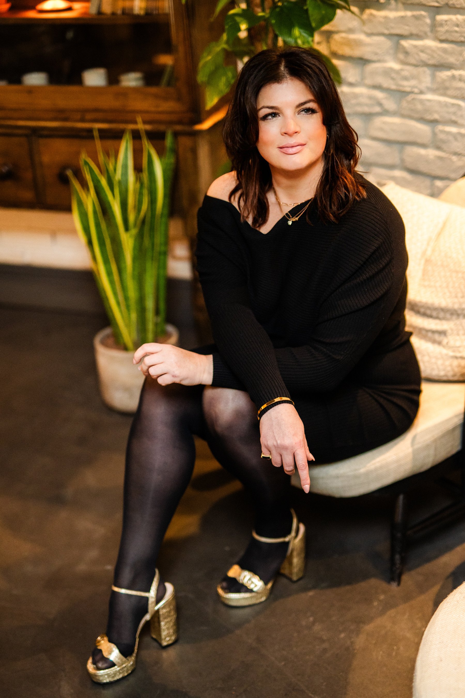A woman with dark hair, wearing a black off-the-shoulder dress, black stockings, and gold high-heeled shoes, sitting on a beige cushioned bench in a cozy, well-lit room with a brick wall, green plants, and wooden furniture.