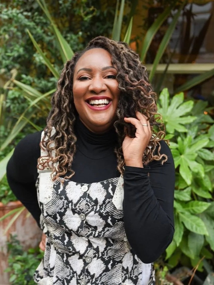 A woman with curly hair and a big smile, wearing a black turtleneck and a snake-patterned overalls, standing outdoors with green plants in the background.