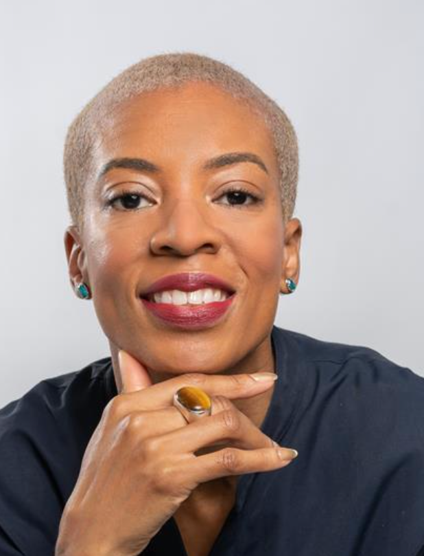 Close-up portrait of an African American woman with short blonde hair, smiling, wearing turquoise earrings, a dark top, and a ring on her finger, resting her chin on her hand against a light background.