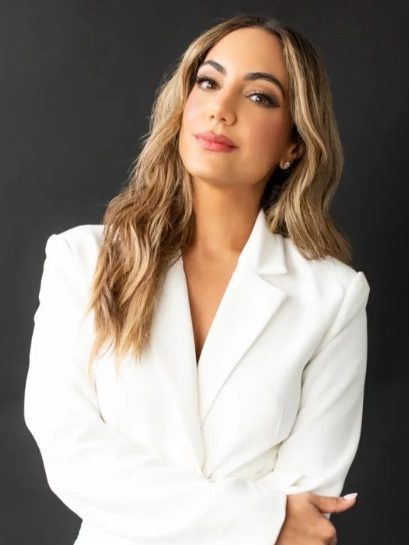 A woman with long wavy blond hair wearing a white blazer, standing against a dark gray background.