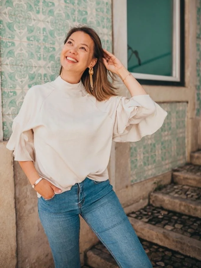 A woman with shoulder-length hair smiling and posing with one hand touching her hair, standing outdoors near stairs and tiled walls.