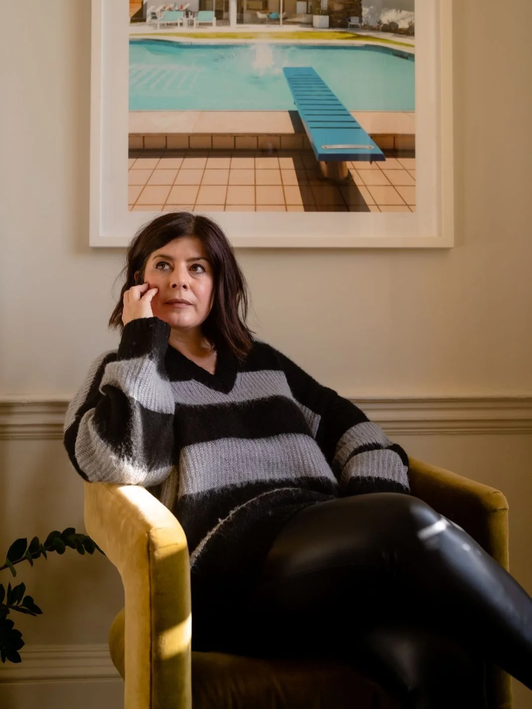 A woman with dark hair sitting on a yellow armchair, resting her head on her hand, in front of a framed picture of a swimming pool.