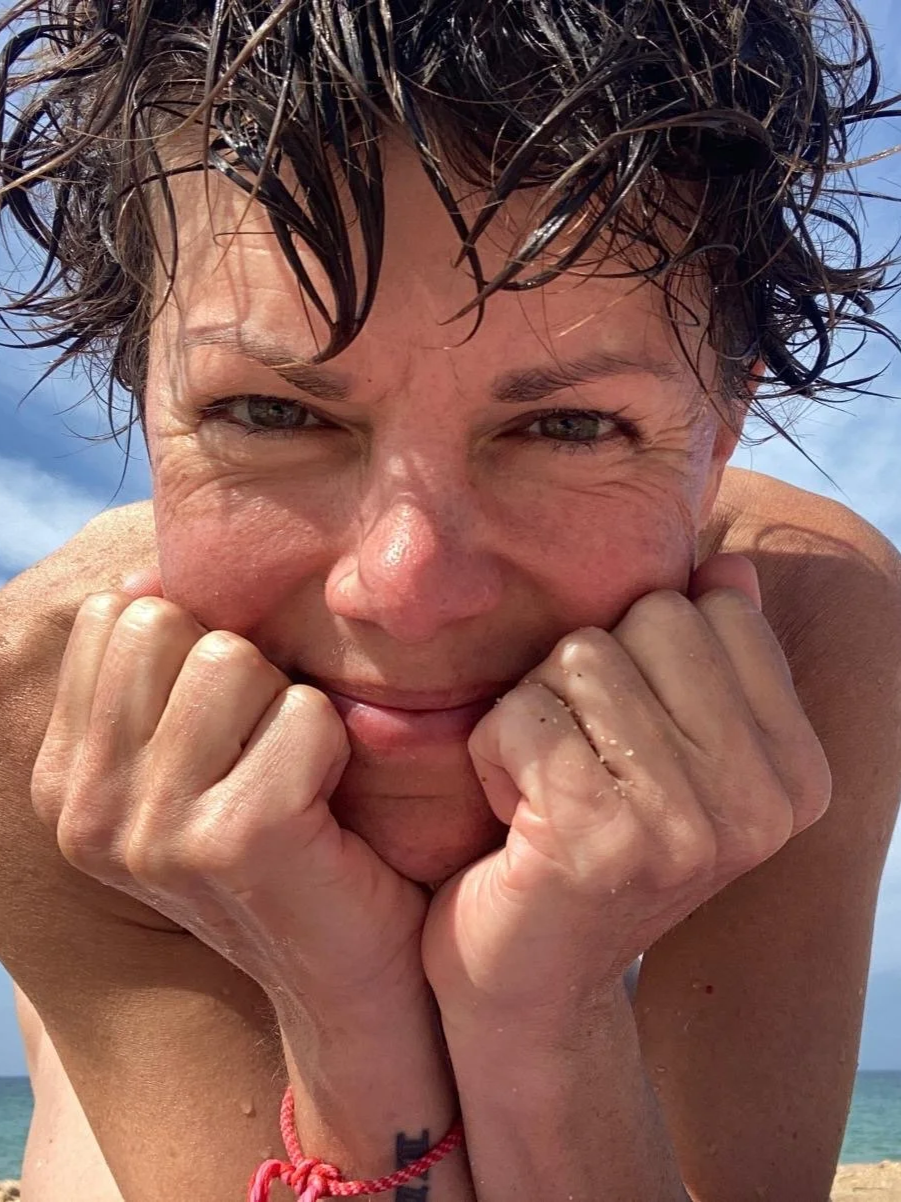 Close-up of a smiling woman with wet hair, resting her chin on her fists, on a beach with a blue sky in the background.