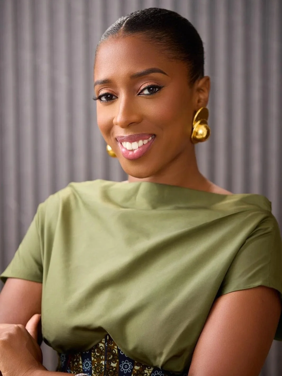 A woman smiling, wearing gold earrings and an olive green top, standing in front of a gray curtain.