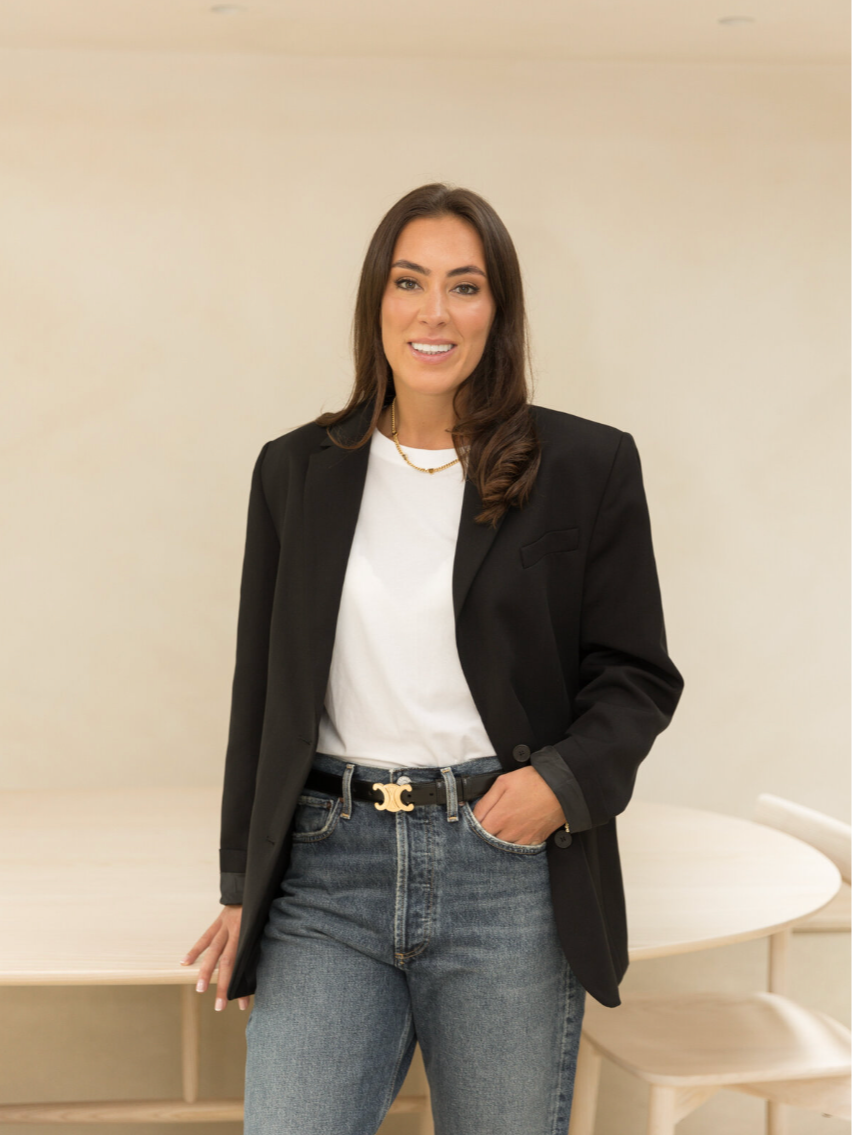A woman with long dark hair, wearing a black blazer, white t-shirt, and jeans, standing indoors near a table and smiling.