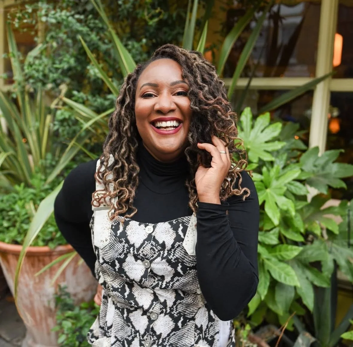 A woman with curly hair wearing a black turtleneck and a snakeskin-patterned overall smiling outdoors with green plants in the background.