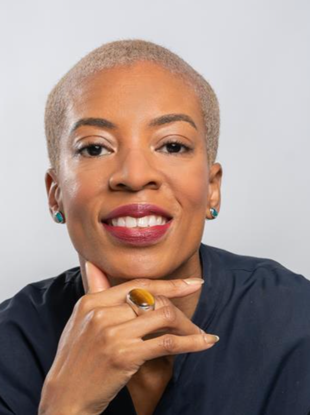 Portrait of a confident woman with short platinum blonde hair, smiling, wearing a dark blouse, teal earrings, and a large yellow ring, resting her chin on her hand.