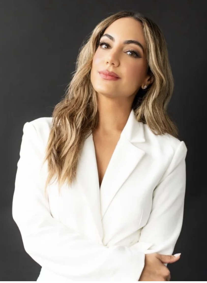 A woman with long wavy hair wearing a white blazer, standing against a dark gray background.