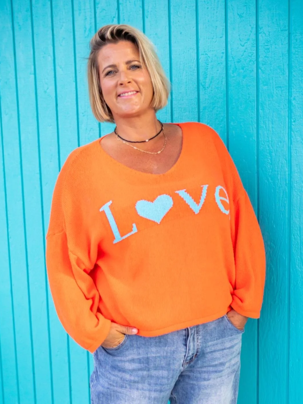 A woman with short blonde hair, smiling, wearing an orange sweater with the word 'LOVE' and a heart symbol on it, standing in front of a blue wooden wall.