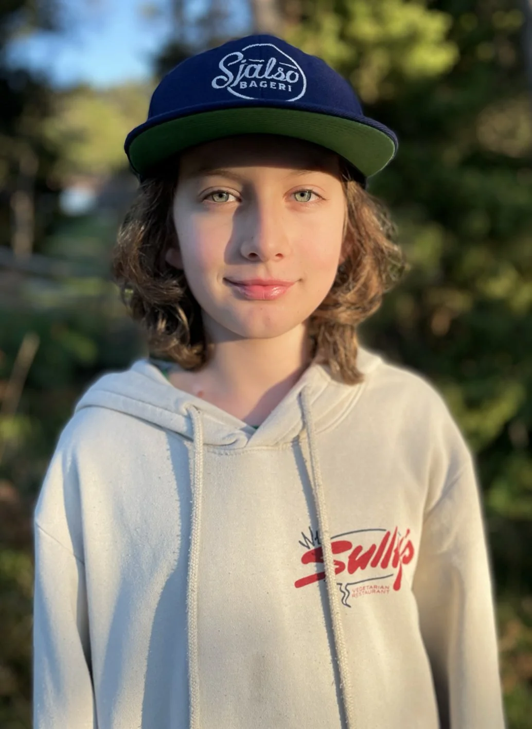 A young person with shoulder-length curly hair, fair skin, and blue eyes, wearing a navy blue and green cap with the words 'Sialso Bageri' and a white hoodie with red and black text, standing outdoors in natural sunlight with a blurred background of