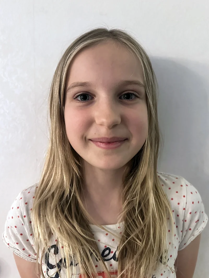 Gotland pictures film casting - A young girl with long blonde hair, blue eyes, and light skin, smiling softly at the camera, wearing a white shirt with red polka dots, standing against a plain white wall.