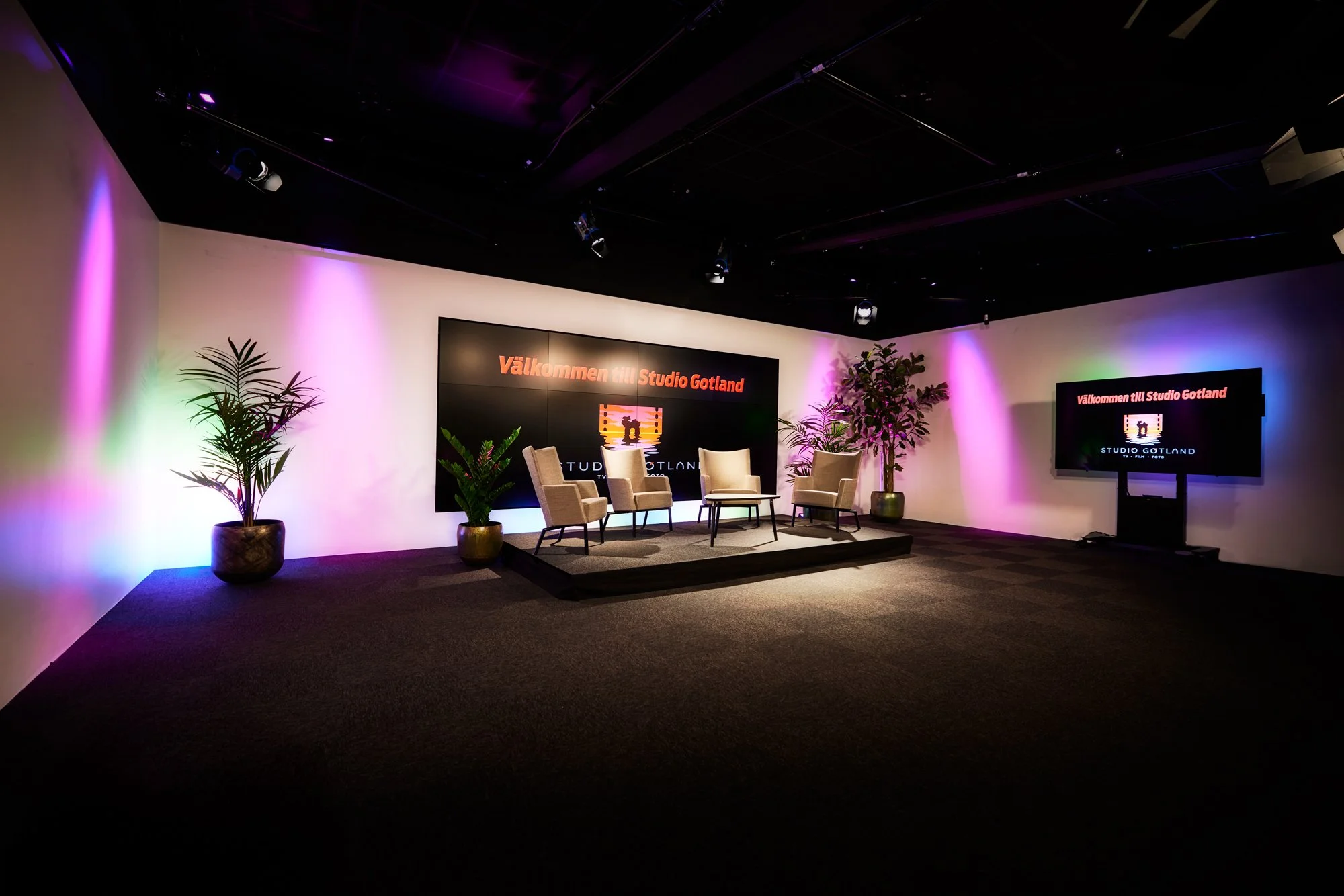 A stage with four chairs and potted plants, two on each side, in a room with purple and green lighting. A large screen in the background says 'Välkommen till Studio Gotland' and 'Studio Gotland' below a logo.
