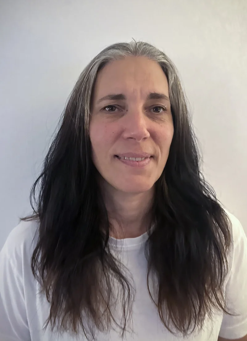 Gotland pictures film casting - A woman with long, dark wavy hair and light skin, wearing a white shirt, standing against a neutral background.