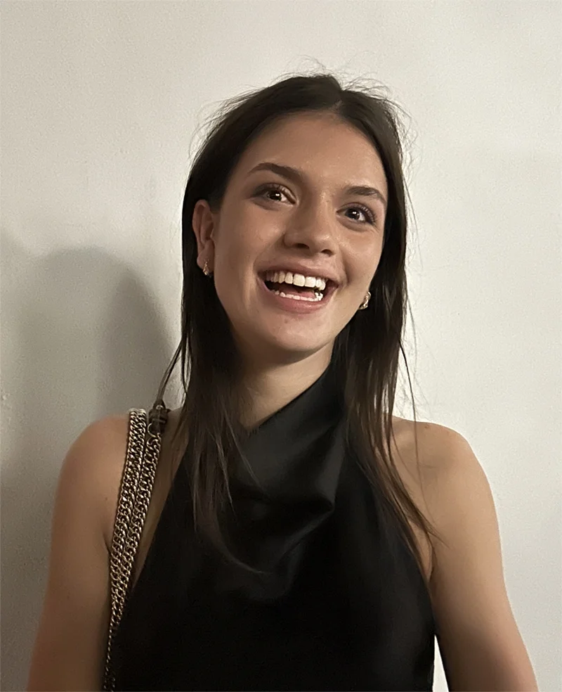 A young woman with brown hair and earrings smiling at the camera, wearing a black sleeveless top and a gold chain strap bag, standing against a plain white wall.
