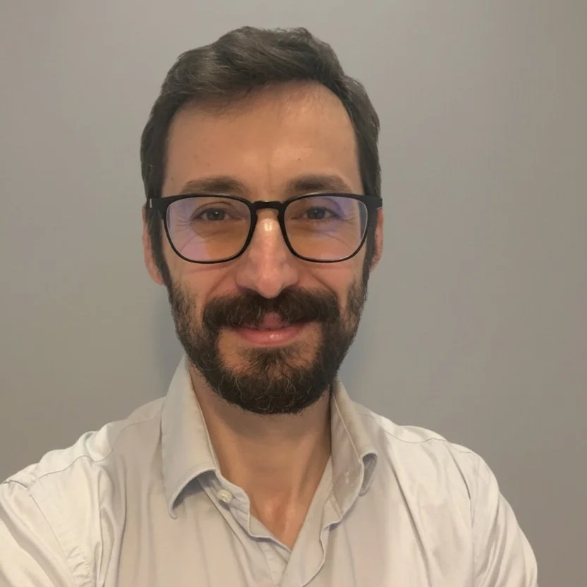 A man with glasses and a beard smiling at the camera, wearing a light-colored shirt, standing against a plain background.