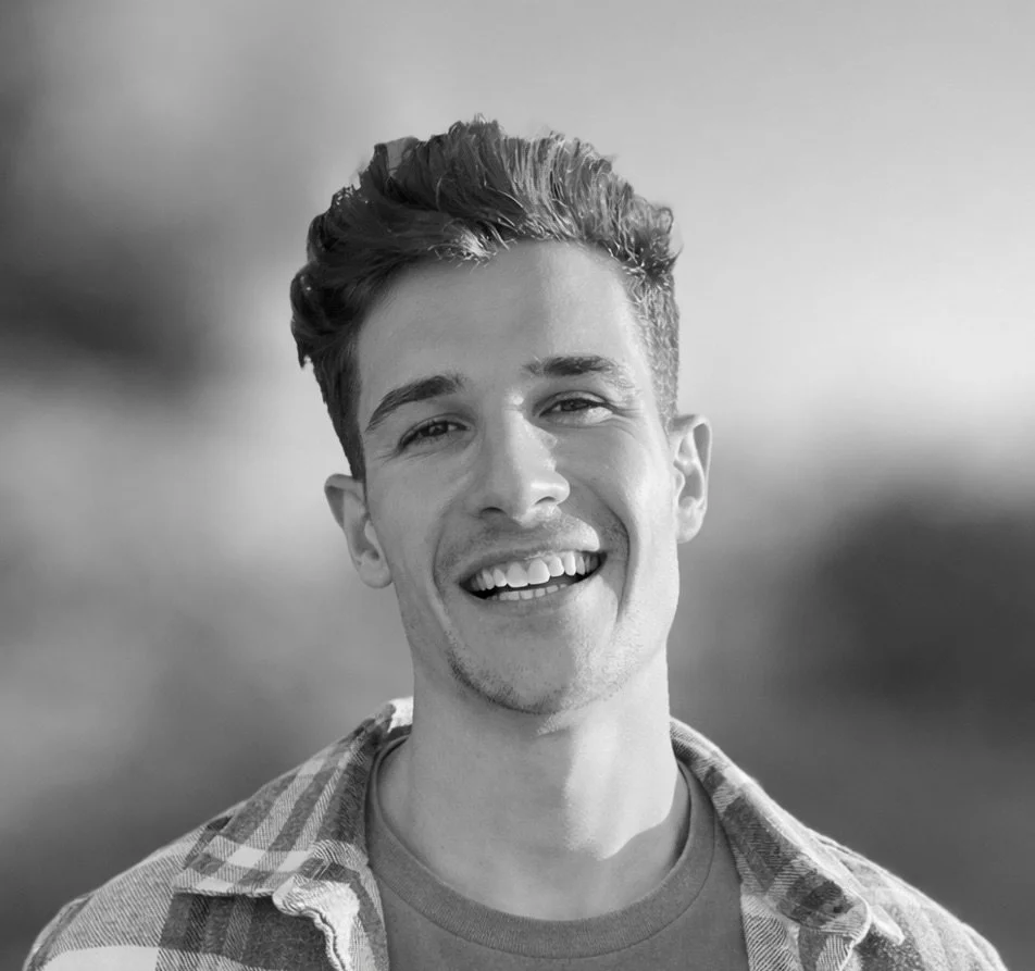 A young man with short curly brown hair wearing clear glasses and a black zip-up jacket, looking at the camera with a slight smile against a plain gray background.