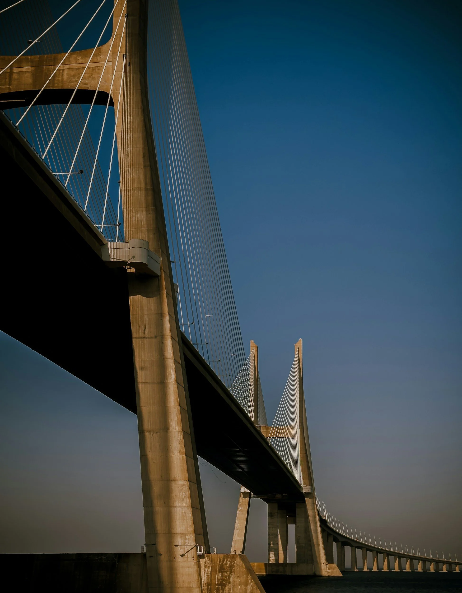 A modern cable-stayed bridge with tall towers and suspension cables, viewed from below against a clear blue sky.