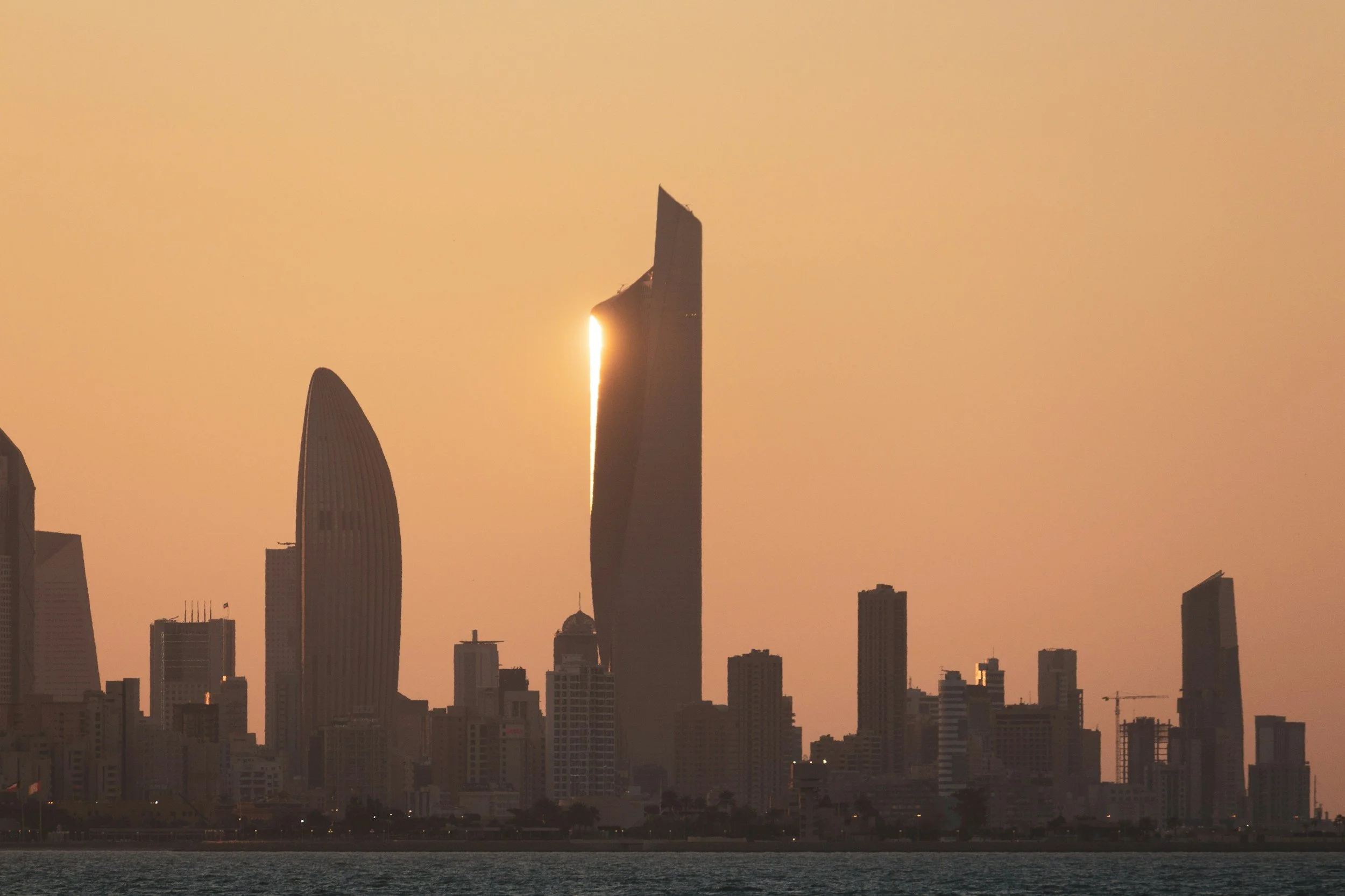 Sunset over a city skyline with tall modern skyscrapers, including a boat-shaped building, reflecting sunlight behind the buildings, with water in the foreground.