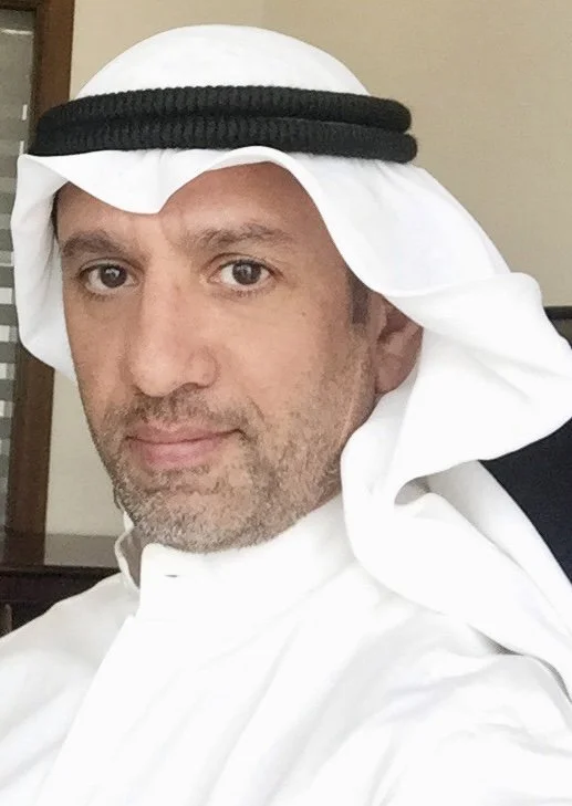 A man wearing traditional Middle Eastern attire, including a white kandura and a white ghutra with a black agal, taking a selfie indoors.