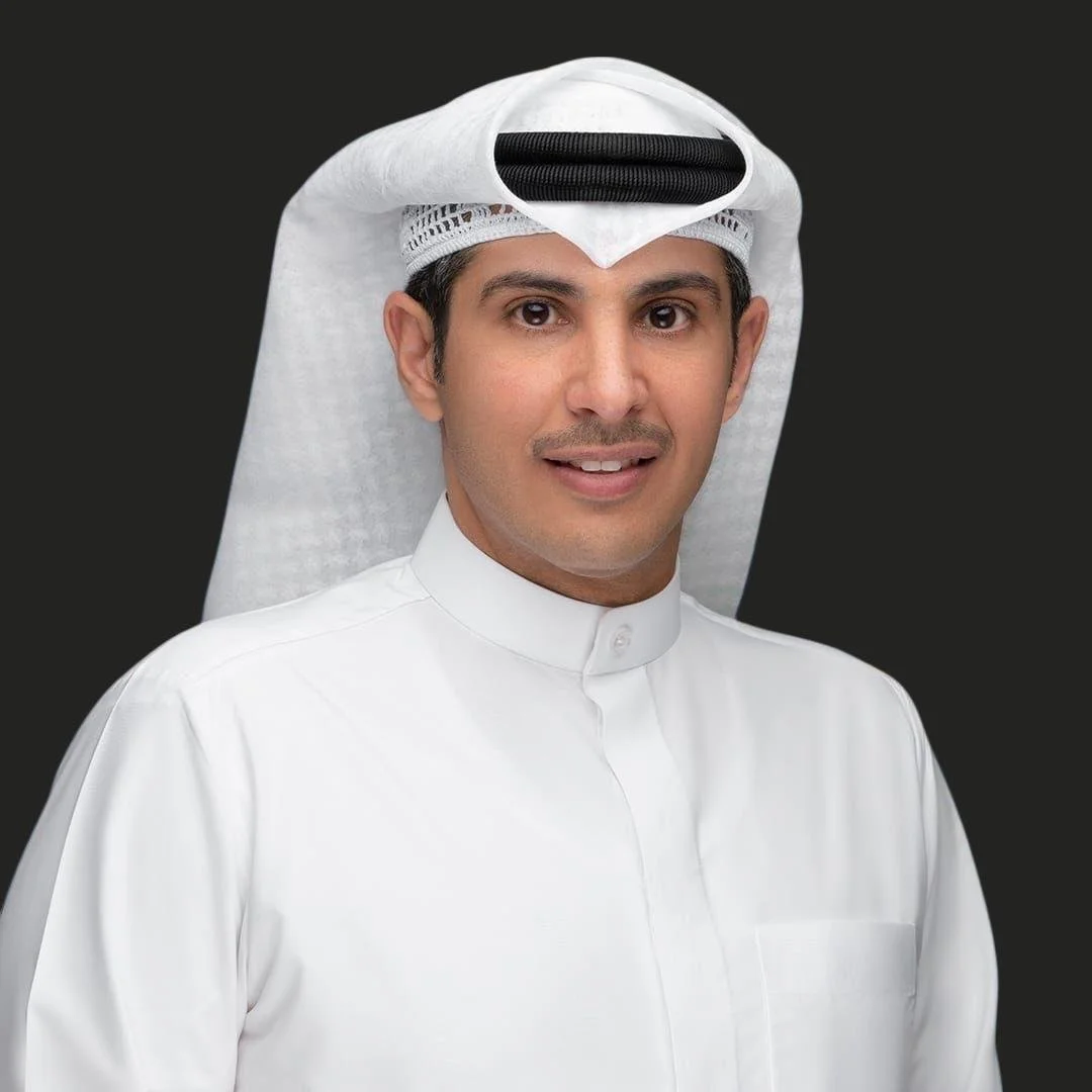 A man wearing traditional Middle Eastern attire, including a white thobe and a white ghutra with a black agal, standing against a dark background.
