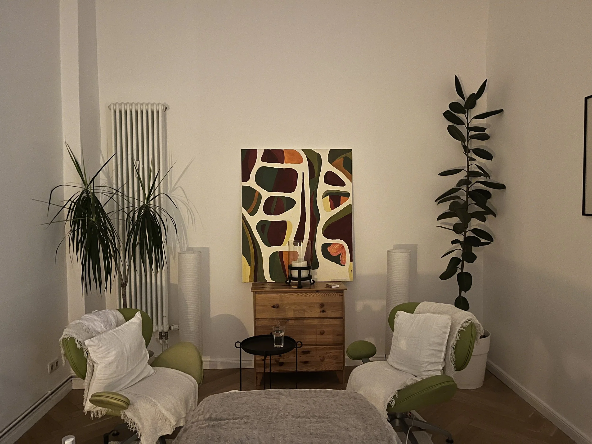 Cozy living room with two green armchairs, each with a white blanket and pillow, facing a small round black table with a glass of water. Behind the table is a wooden chest of drawers with a colorful abstract painting above it. Tall plants are on either side of the painting, one on the left and one on the right, with white lamps behind each plant.