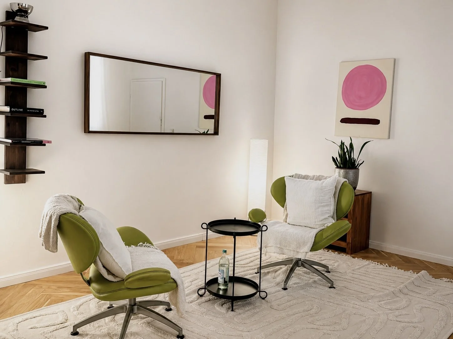 room with two green lounge chairs with white cushions and throws, a beige patterned rug, a small black round table with a bottle of water, a white floor lamp, a potted plant, a large horizontal mirror, a bookshelf, and a pink abstract art piece on the wall.
