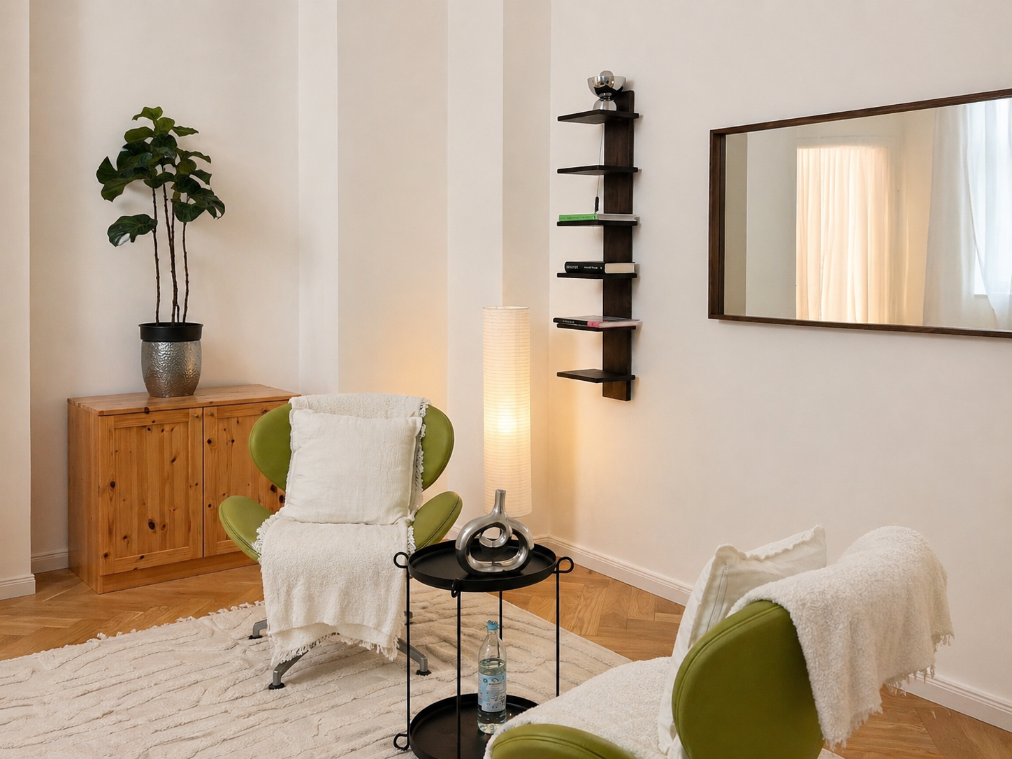 Therapy room with two green armchairs with white throws, a black side table with a decorative silver sculpture and water bottle, a tall floor lamp, a wooden cabinet with a large plant on top, a mirror on the wall, and a modern wall shelf with books.