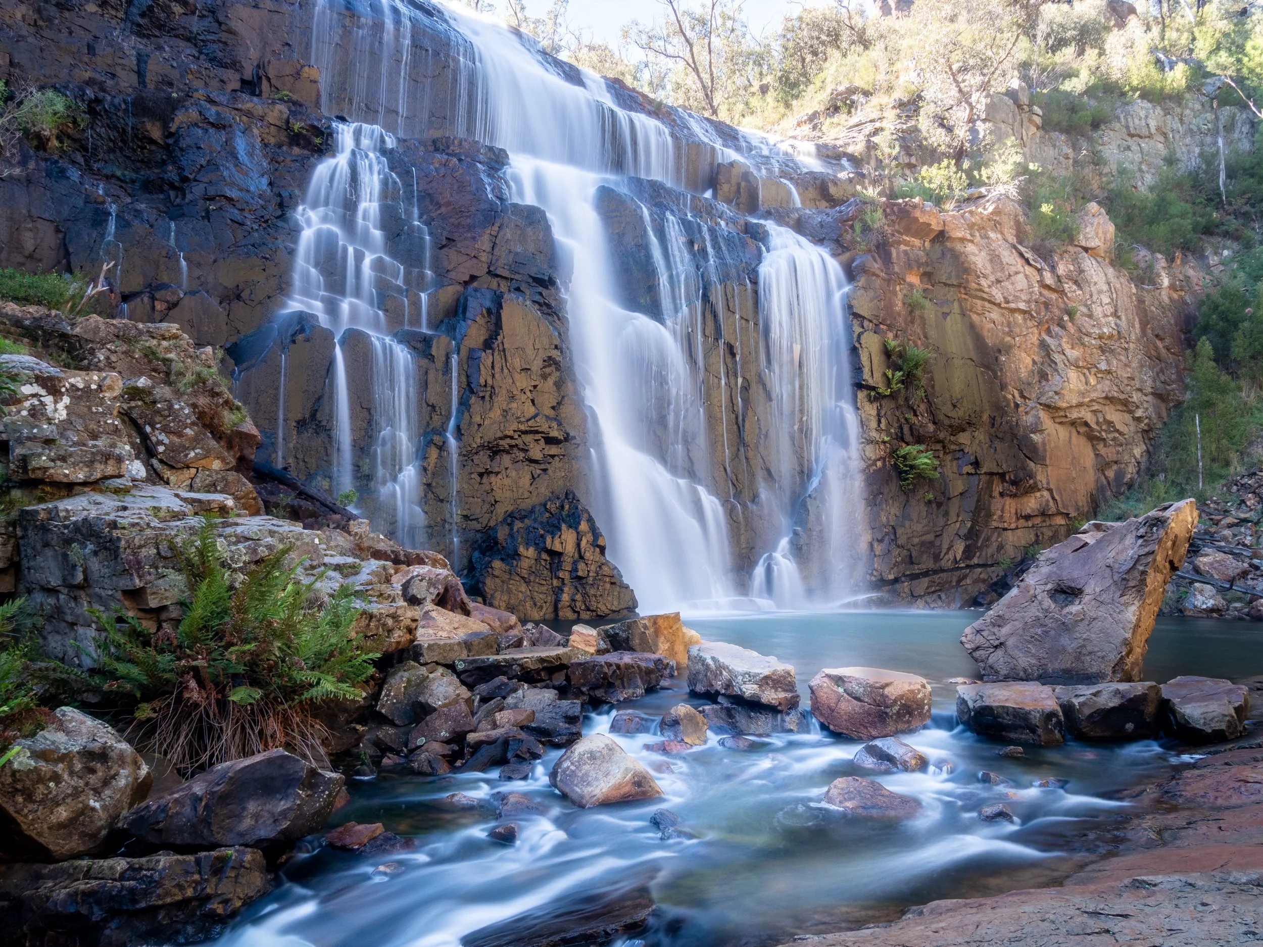 A waterfall flowing down a rocky cliff into a pool below, surrounded by rocks and greenery.