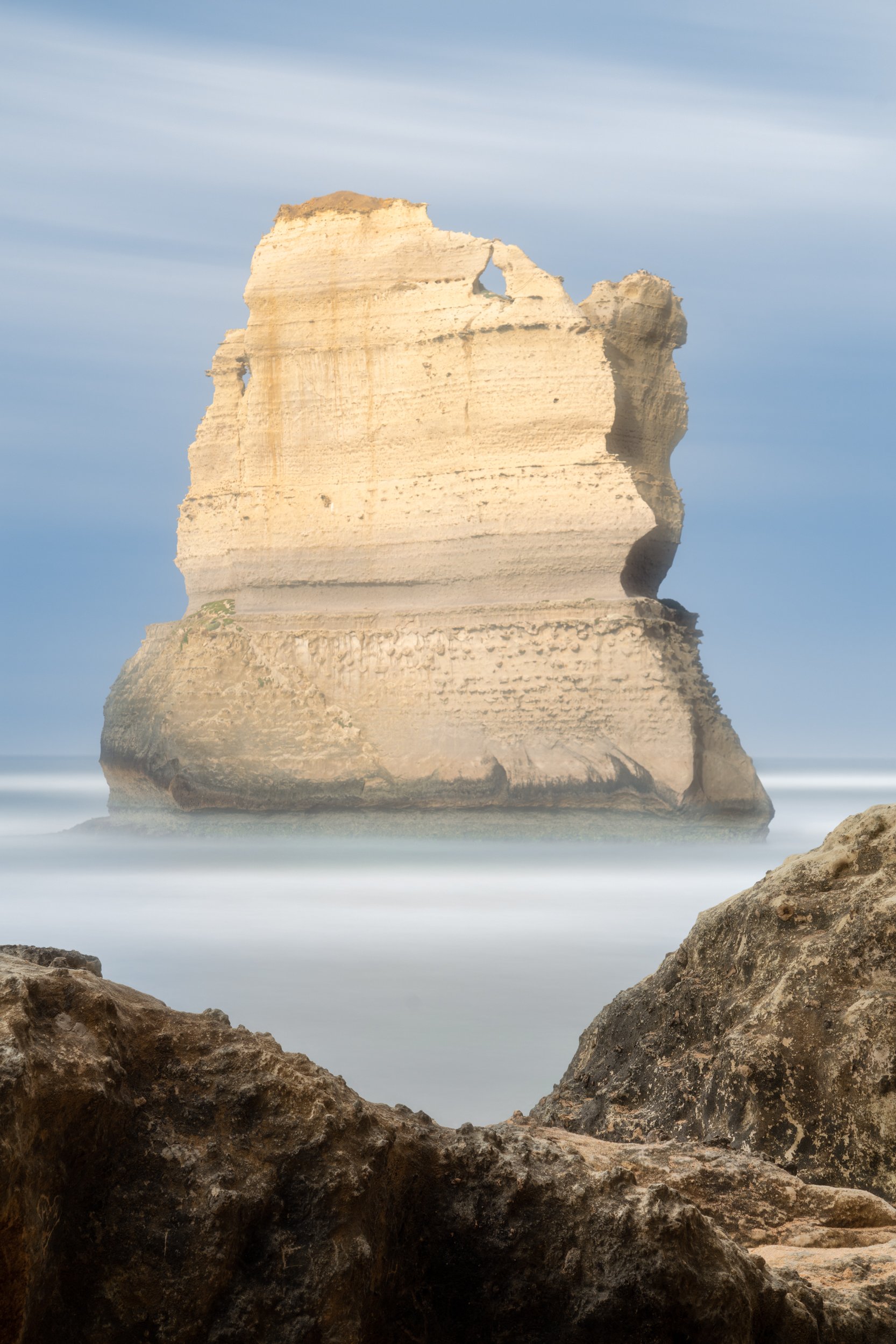 A large sea stack with layered rock formations rises from the ocean, viewed from rocky shoreline, with a cloudy sky in the background.
