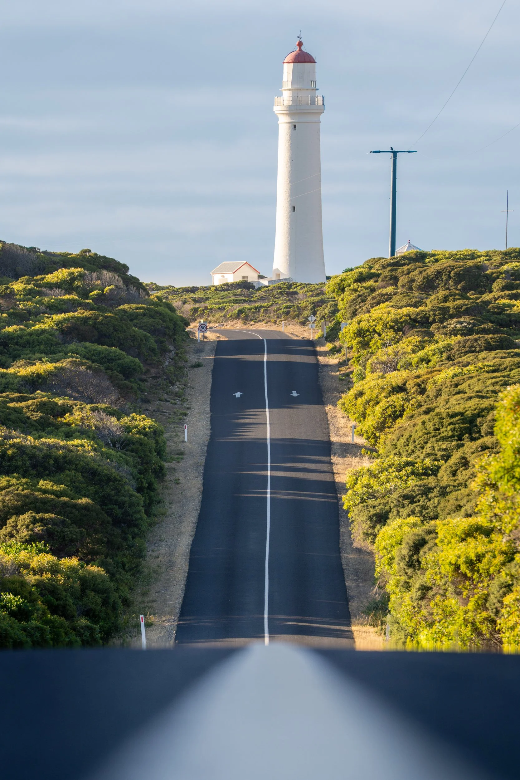 A long straight paved road leading towards a white lighthouse on a hill, with green shrubbery on either side and a partly cloudy sky.
