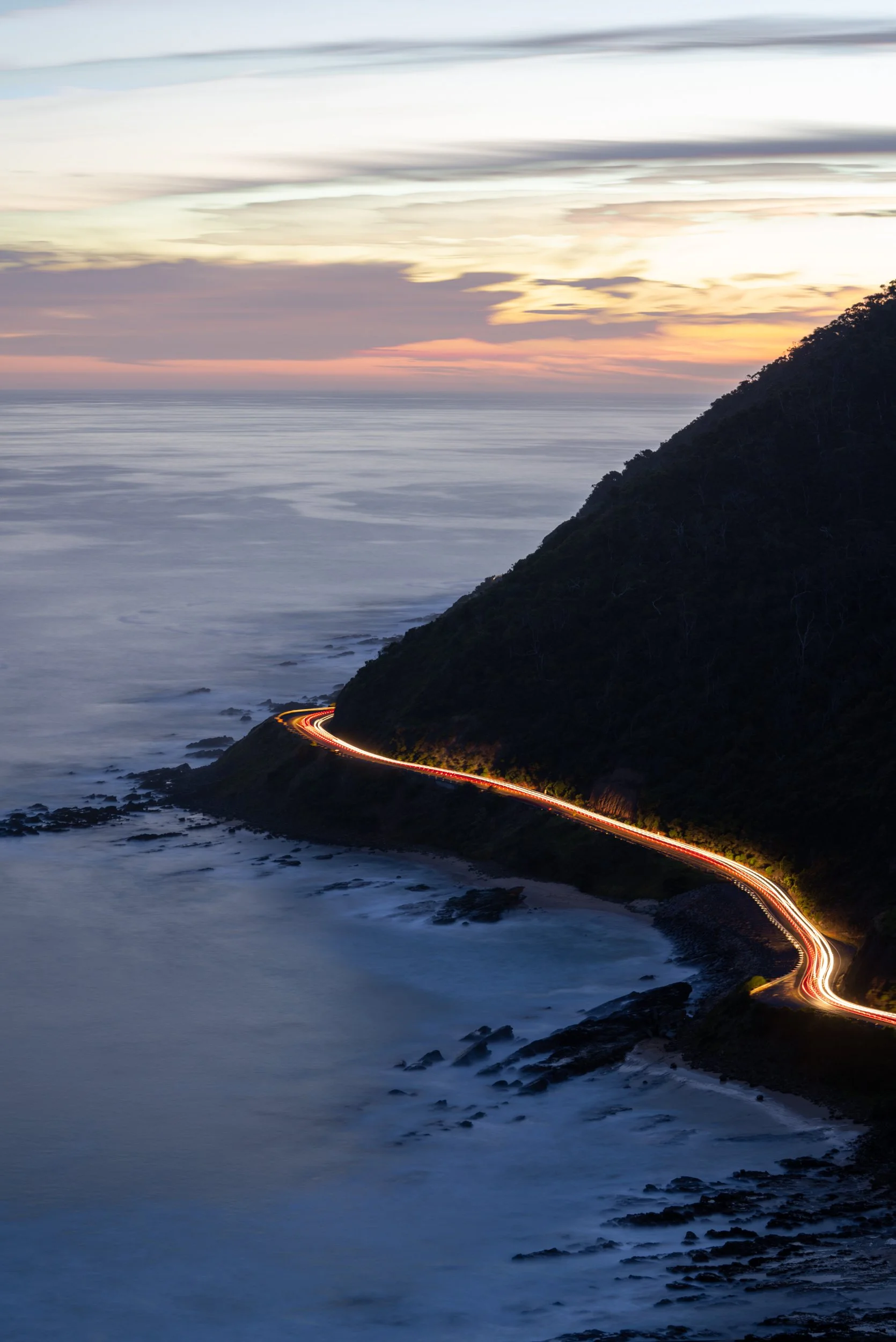 Long exposure photo of a winding coastal mountain road at sunset, with car light streaks and the ocean below.