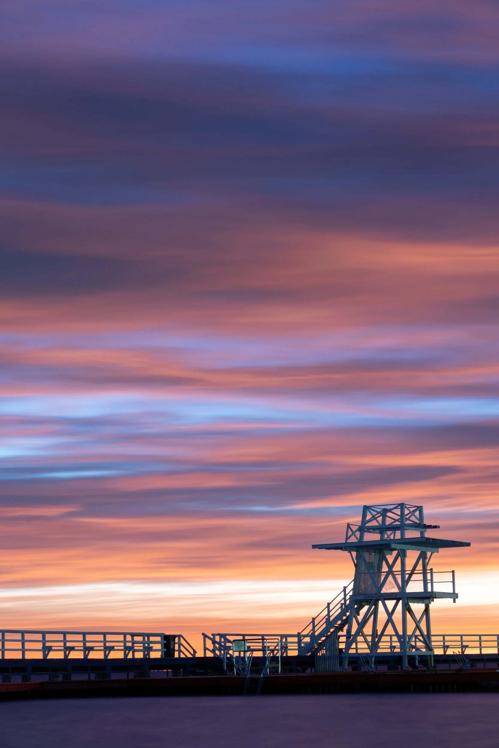 Sunset over a wooden lifeguard tower and pier, with colorful clouds in the sky.