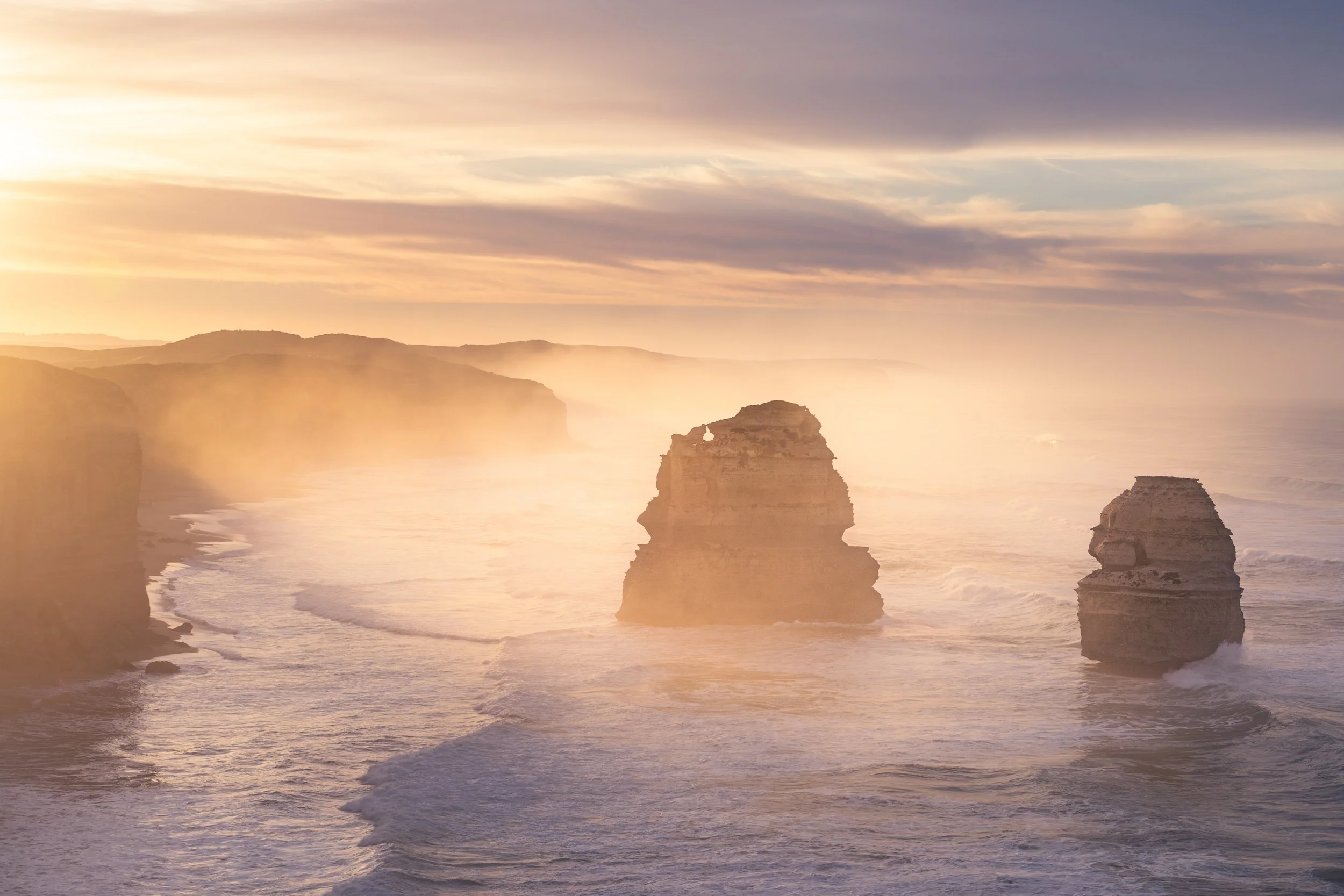 Sunset over the ocean with two large rock formations emerging from the water and cliffs in the background, with a misty atmosphere.