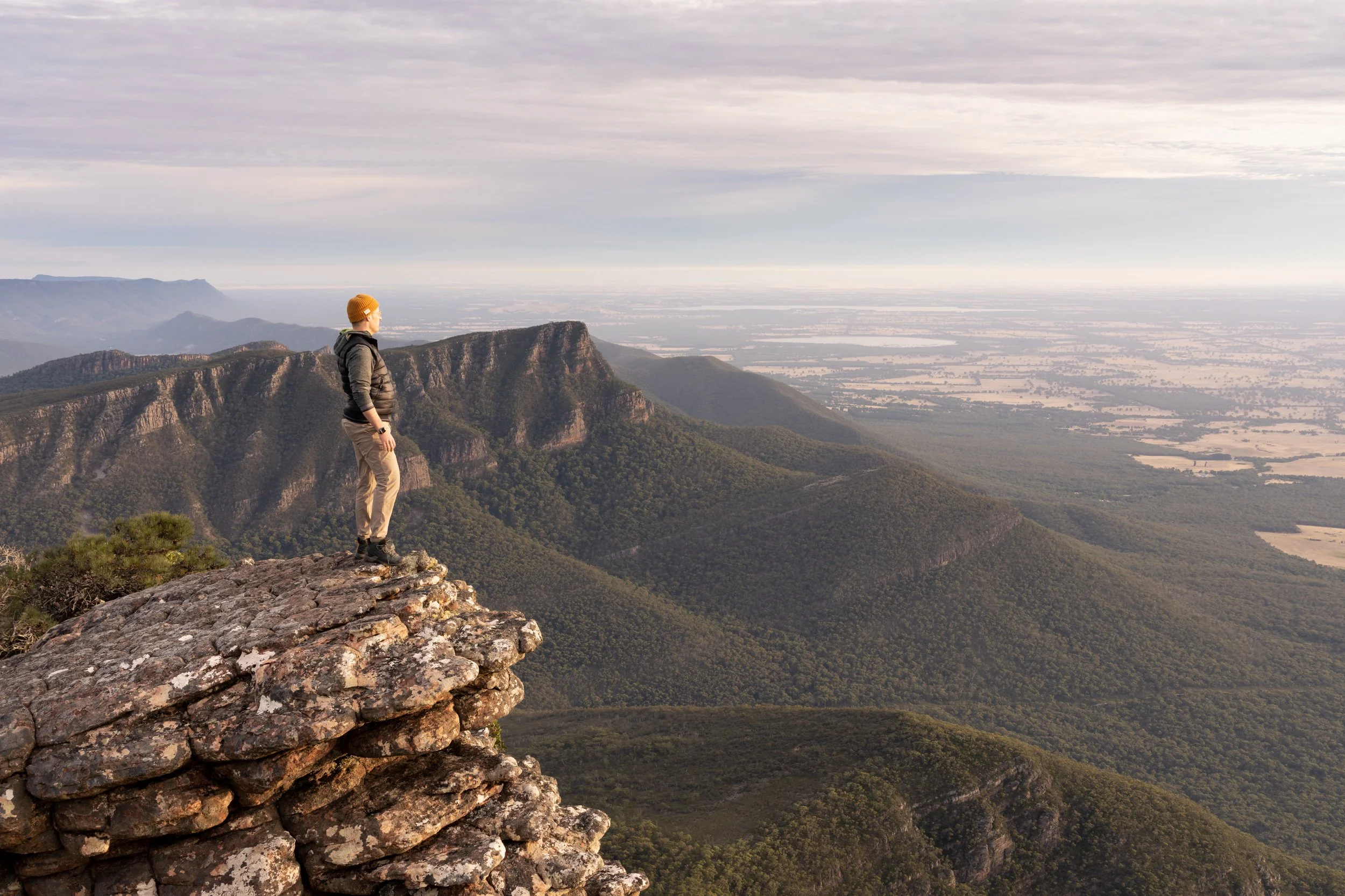 A person standing on a rocky cliff overlooking a scenic valley with mountains and fields in the distance.