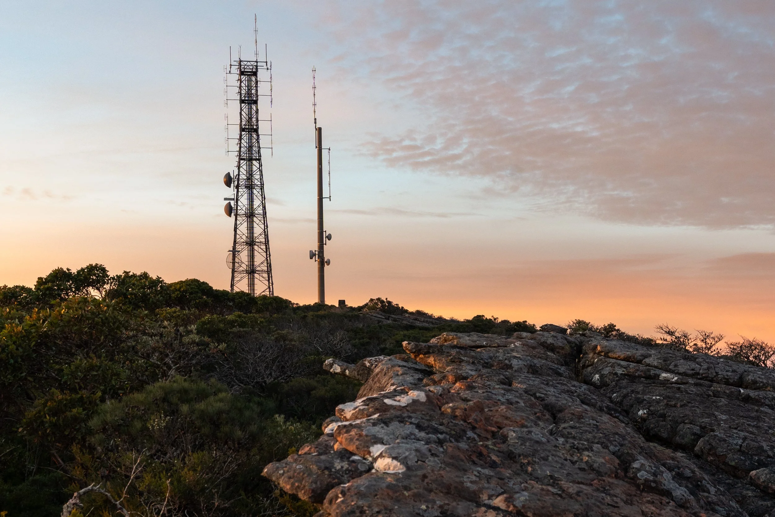 Two radio towers on a hilltop at sunset, with a rocky foreground and sparse vegetation under a partly cloudy sky.