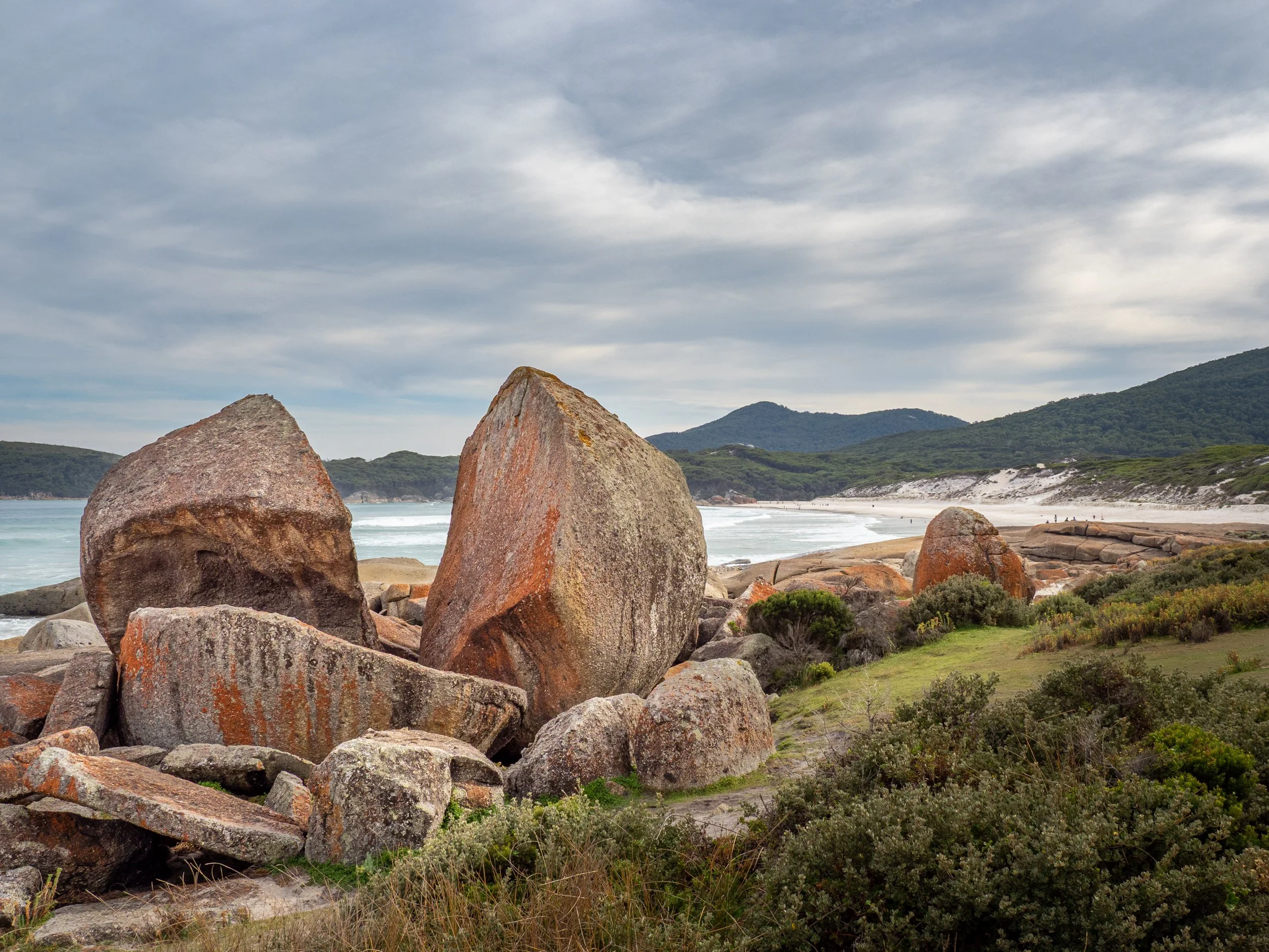 Large rocks on a beach with ocean waves, sandy shoreline, green shrubbery, and hills in the distance under cloudy sky.