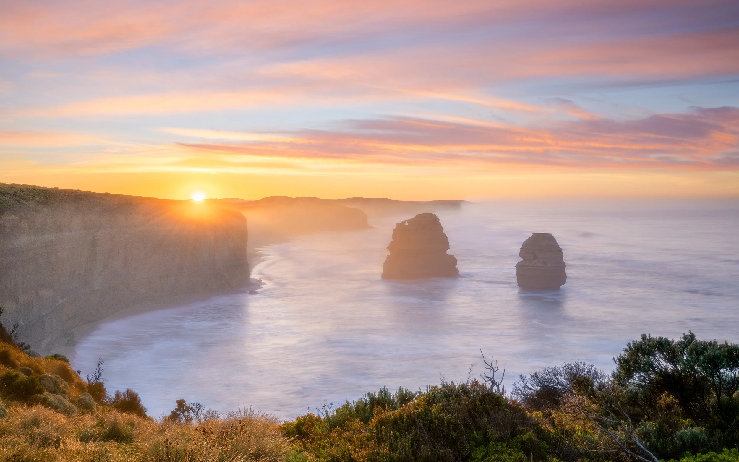 Sunset over the ocean with cliffs and rock formations, overlooking a lush green coastal landscape, mist rising from the water, and colorful sky with pink, orange, and purple hues.