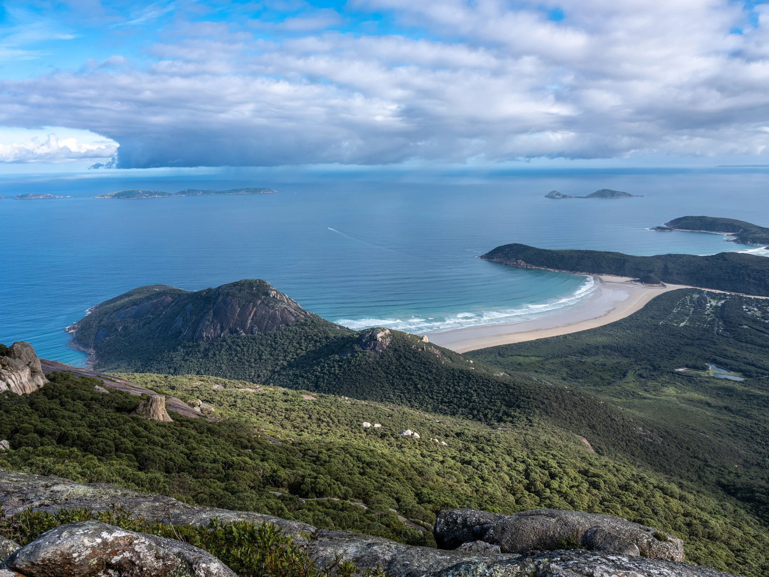 A coastal landscape seen from a high vantage point, featuring green hills, a sandy beach, and calm blue ocean waters with several small islands on the horizon, under partly cloudy skies.