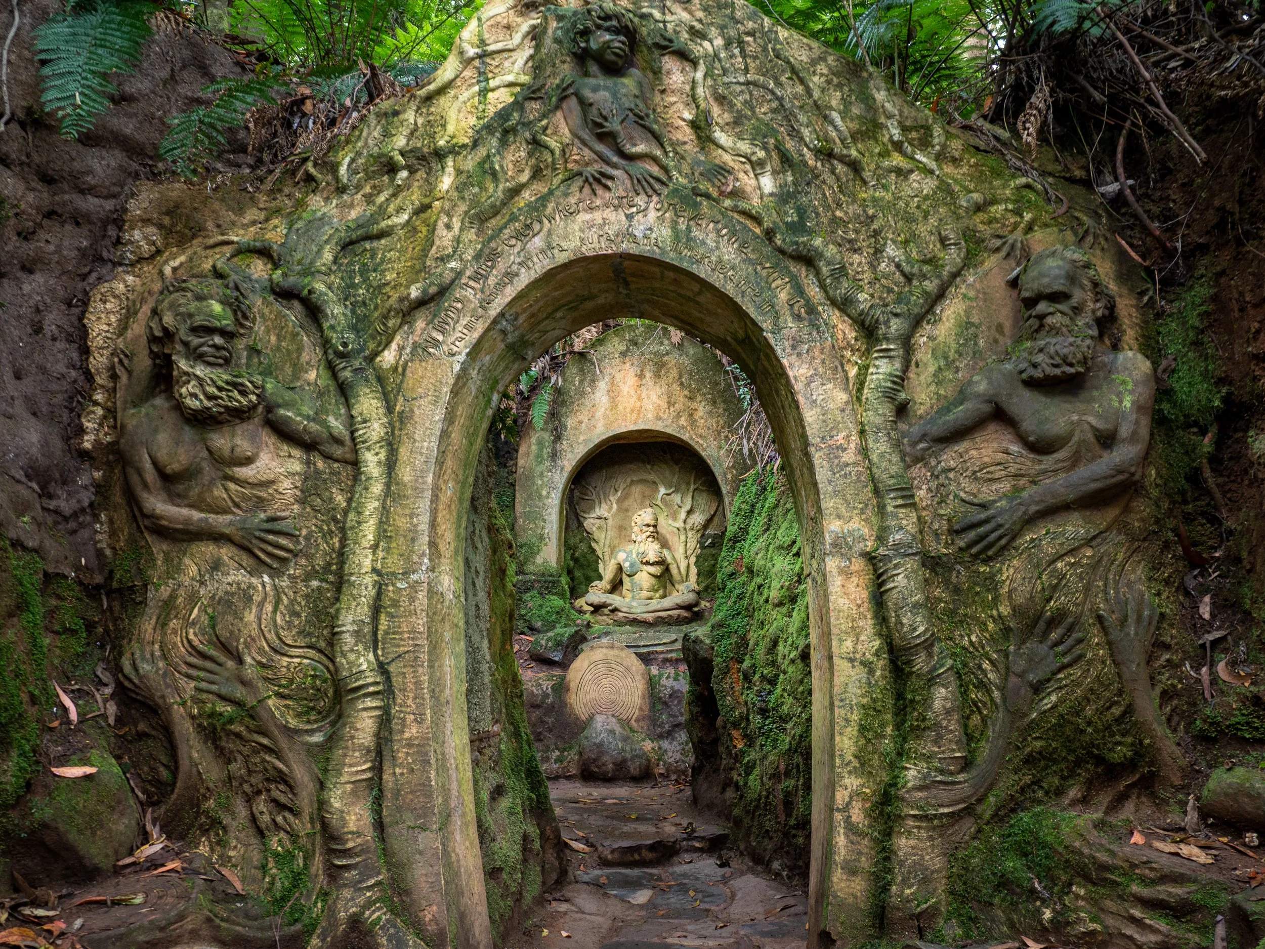 A carved stone archway in a forest with images of biblical figures and a statue of a seated man in meditation at the center.