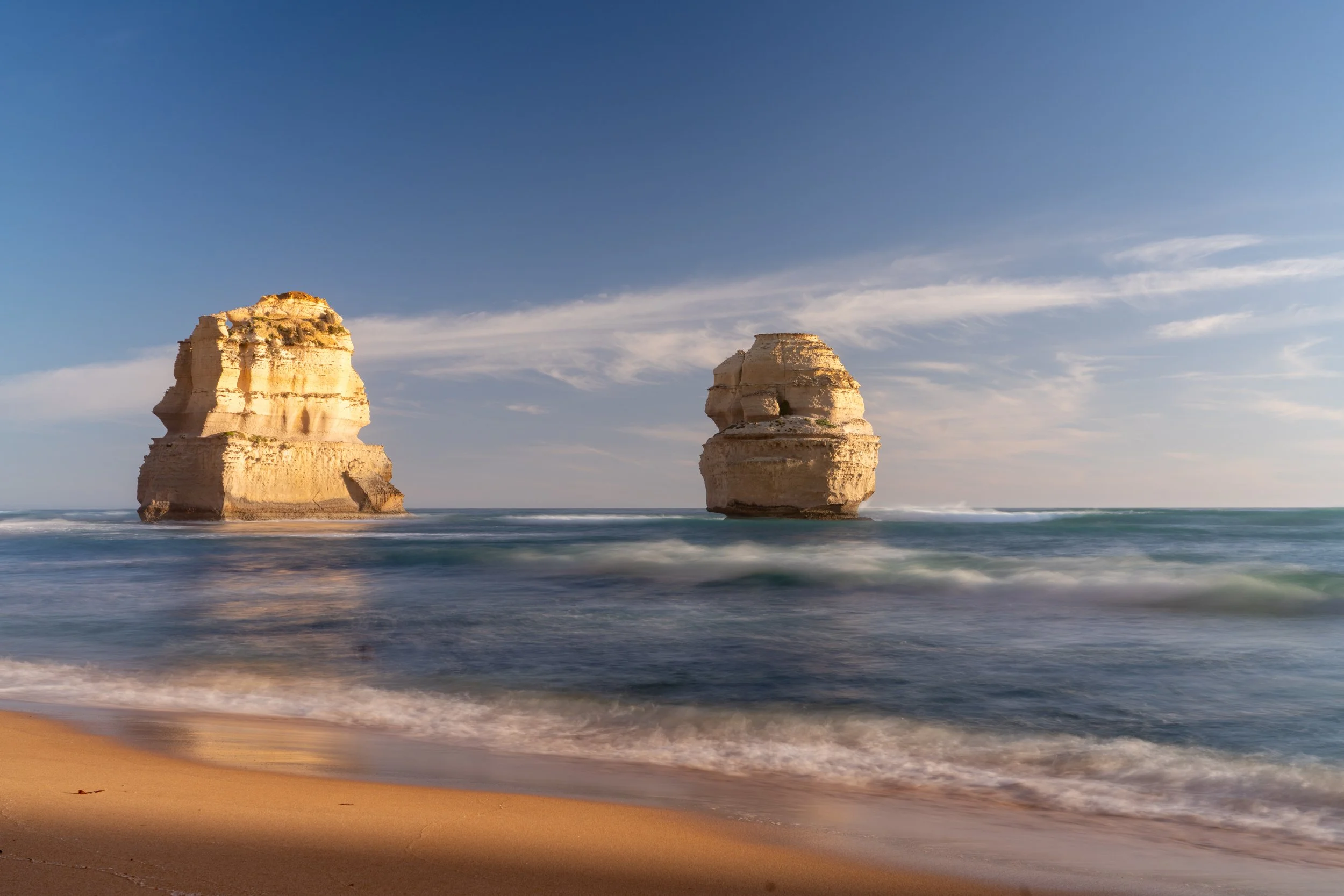 Two large limestone sea stacks in the ocean near a sandy beach under a blue sky with clouds.