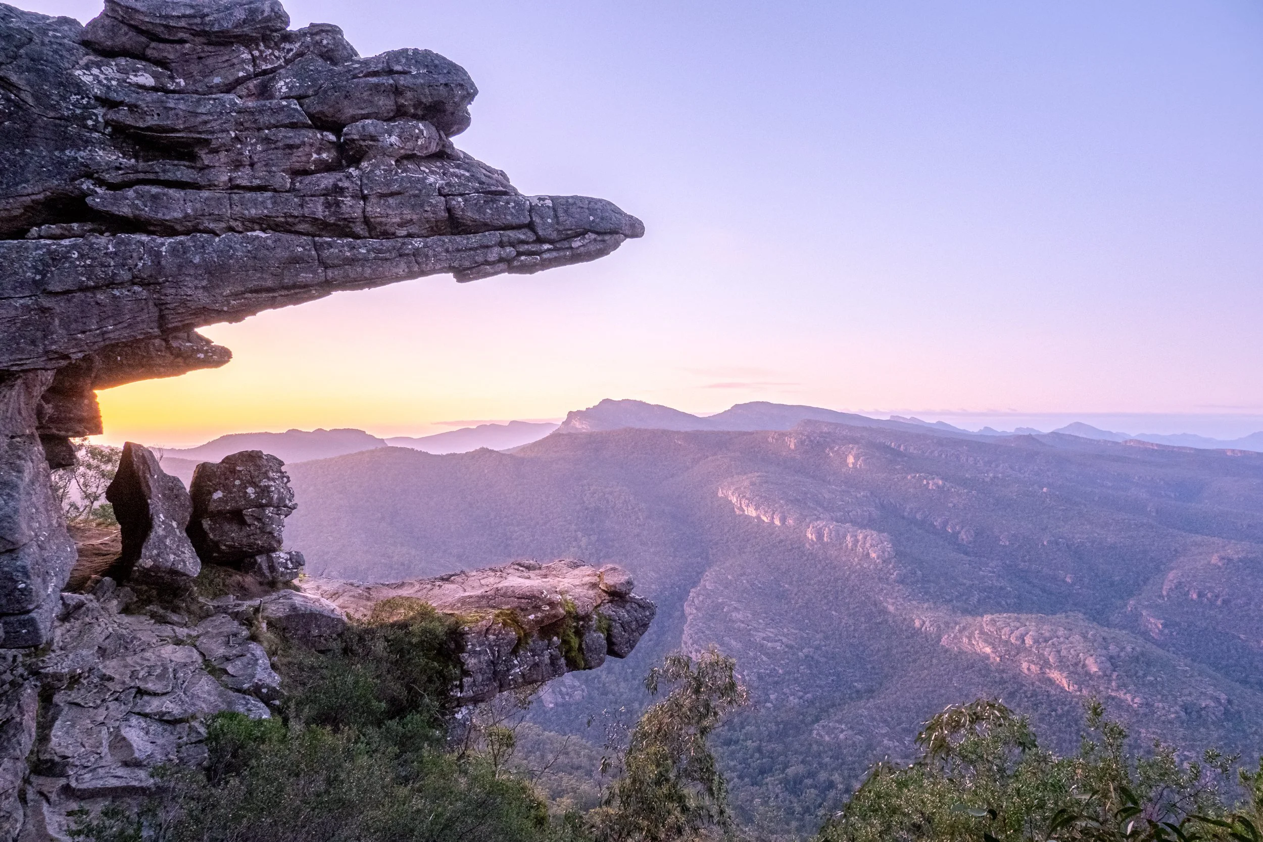 Sunset over a mountain range viewed from a rocky cliff edge with a prominent overhanging rock formation in the foreground.