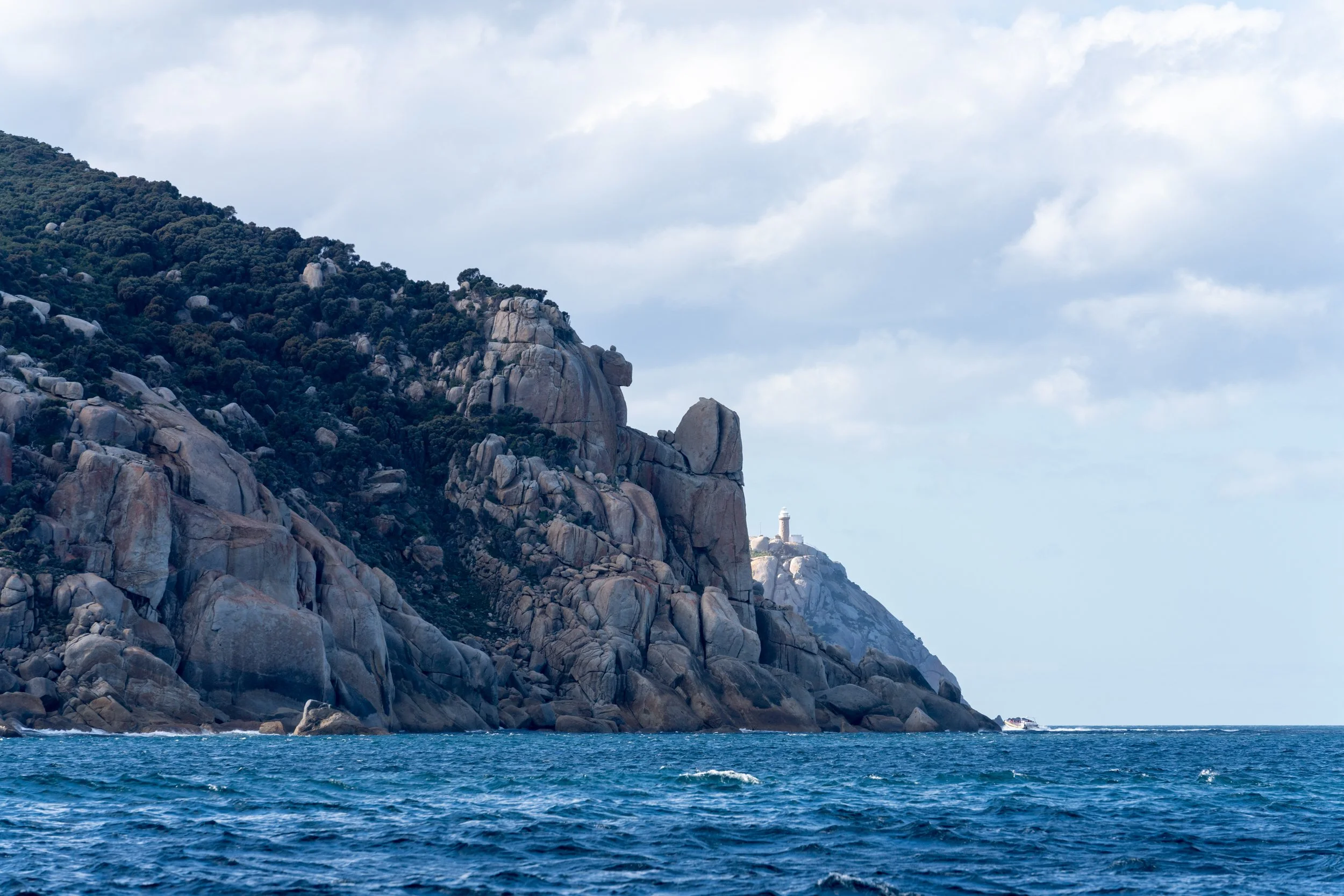 Cliffs along the ocean with a lighthouse visible on the top of the distant hill, partly cloudy sky.