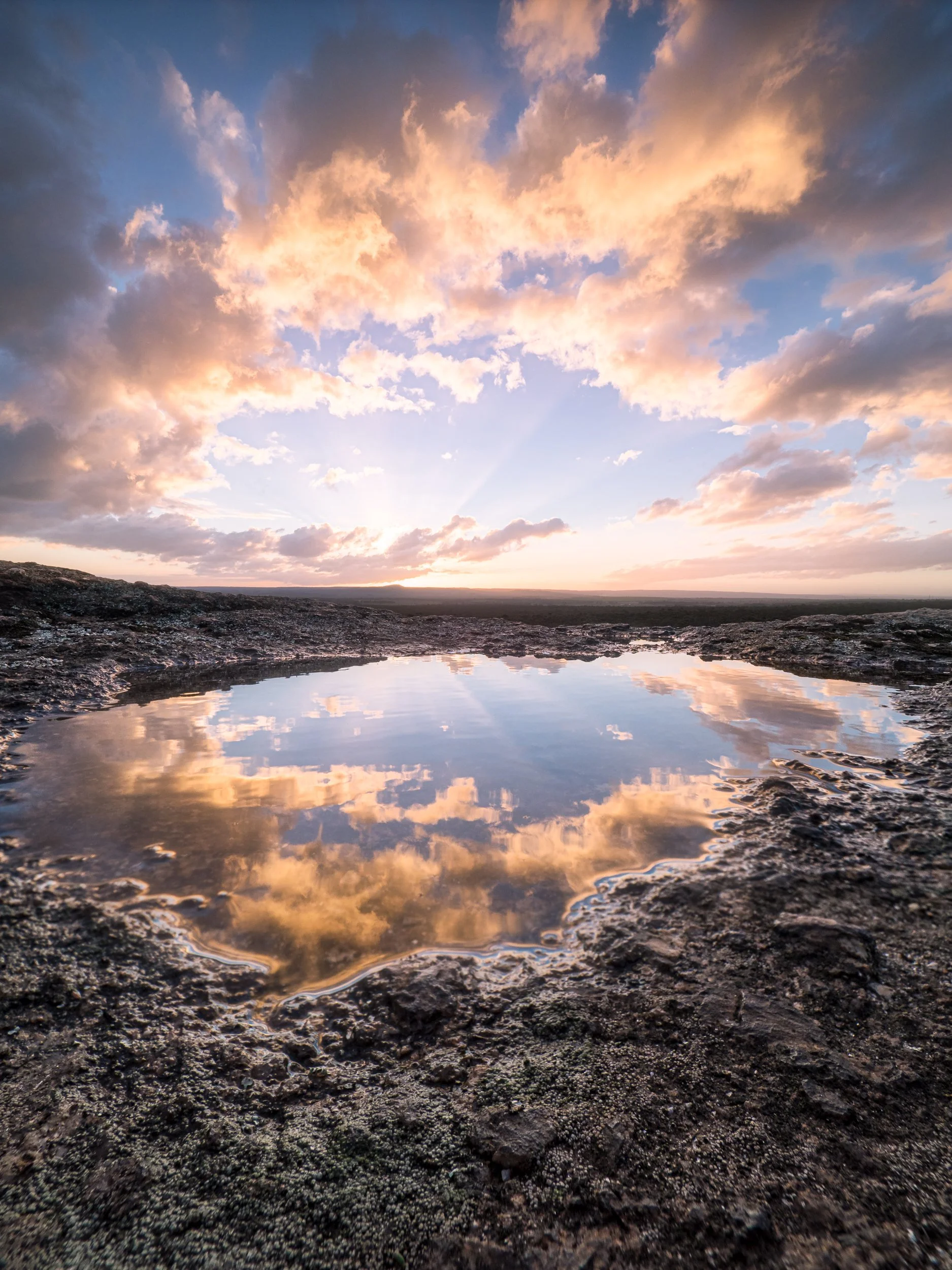 Sunset sky with clouds reflected in a shallow pool on rocky terrain.