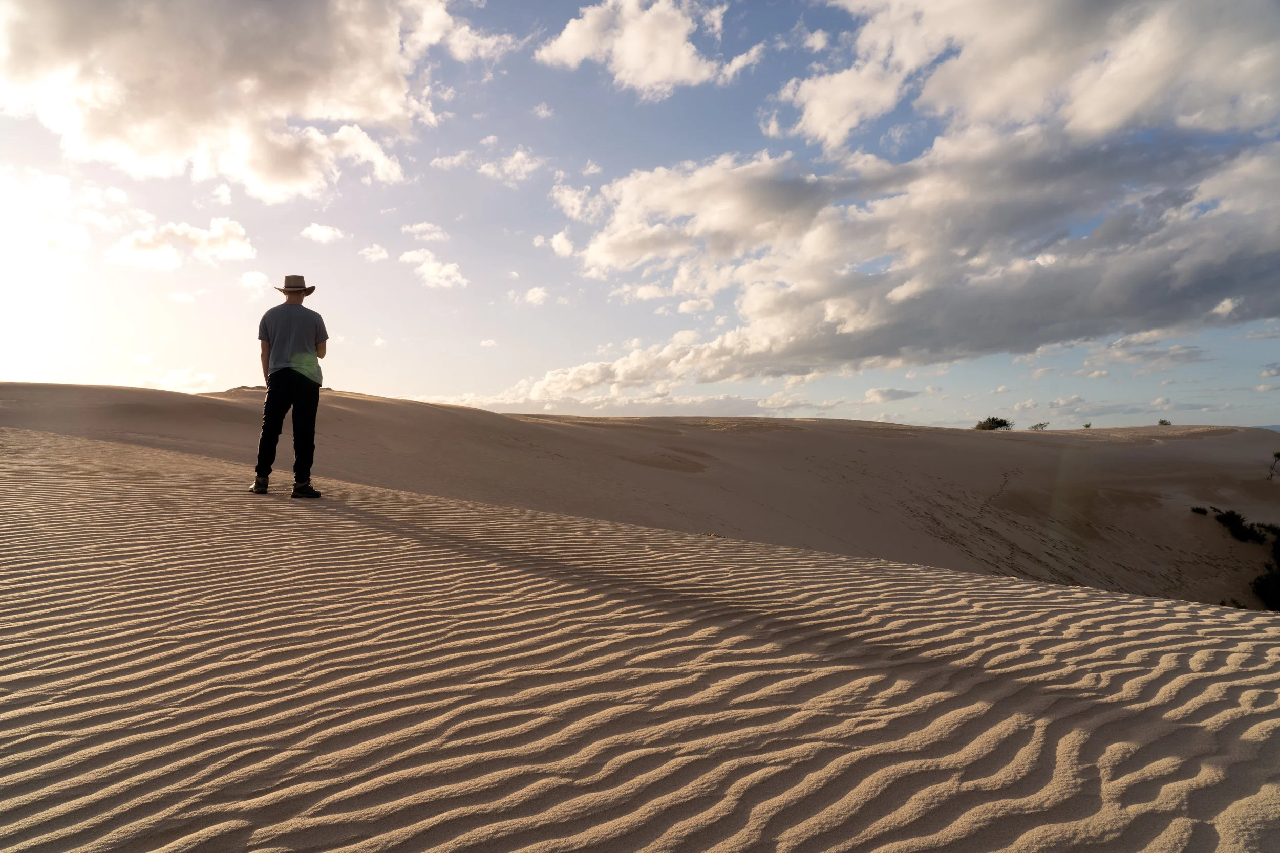 Person wearing a hat standing alone on sandy dunes under a partly cloudy sky during sunset or sunrise.