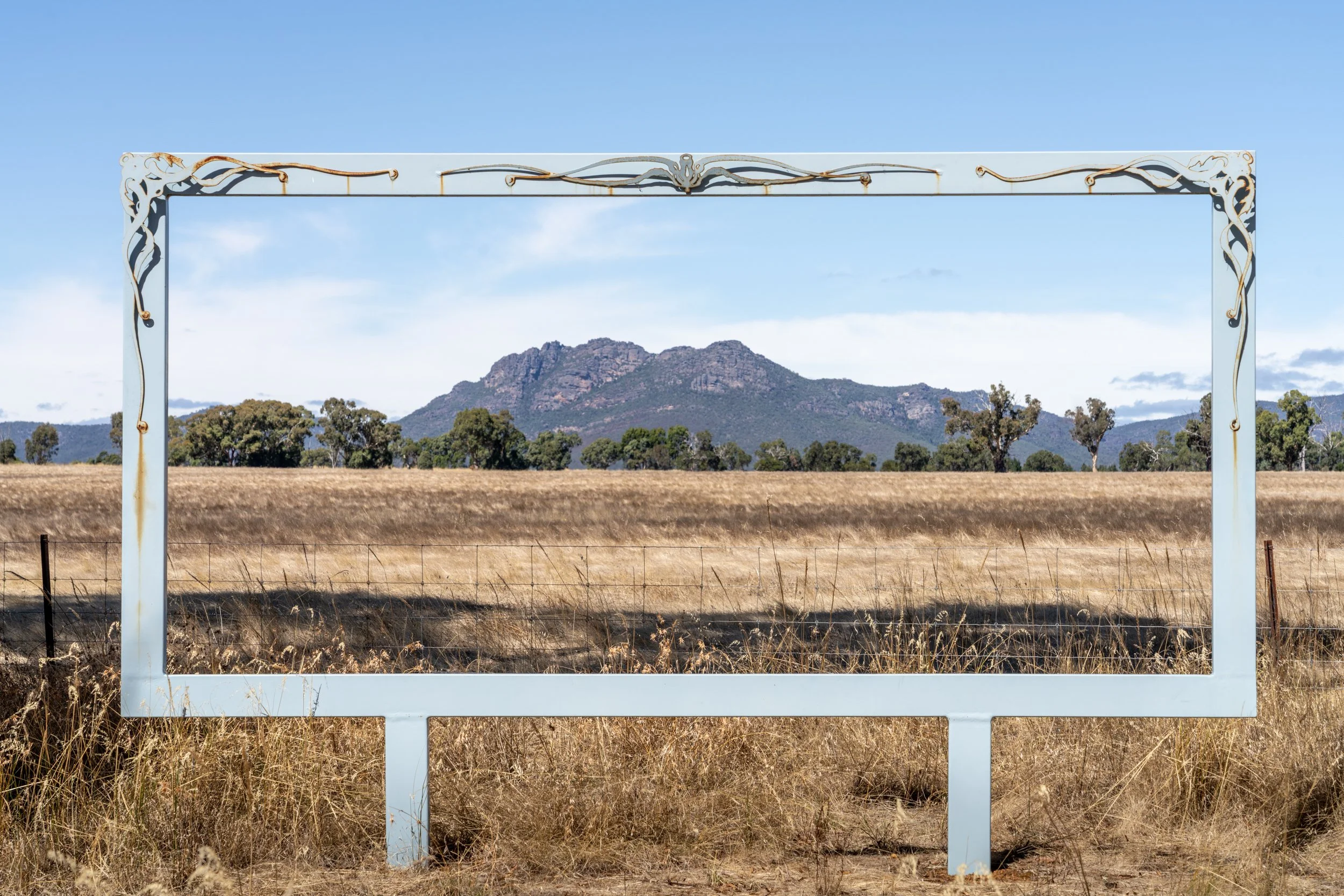 Empty decorative metal frame in an open field with dry grass, trees, and a mountain in the background under a blue sky.
