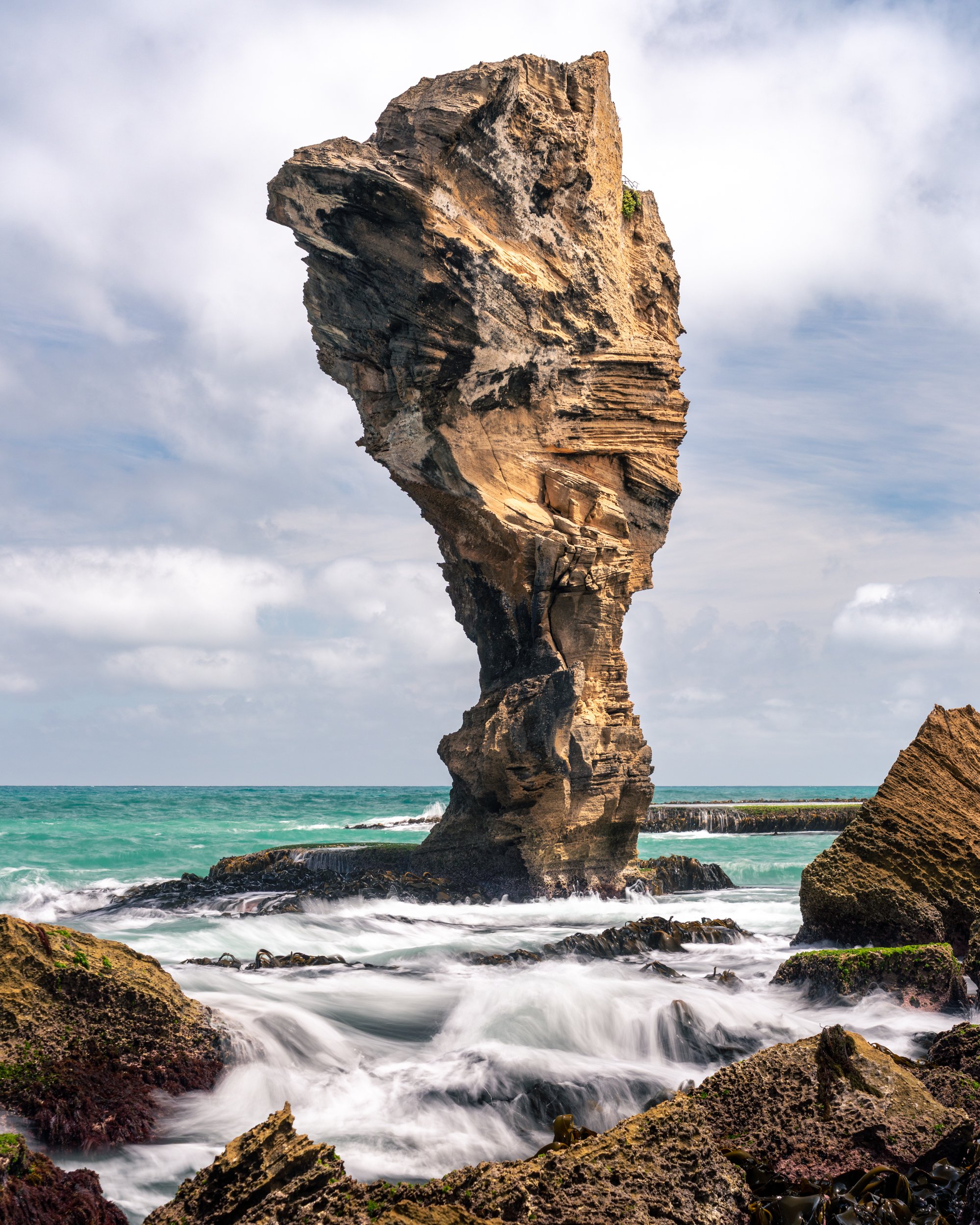 Large rock formation resembling a human face next to the ocean, with waves crashing on rocks at the shore.