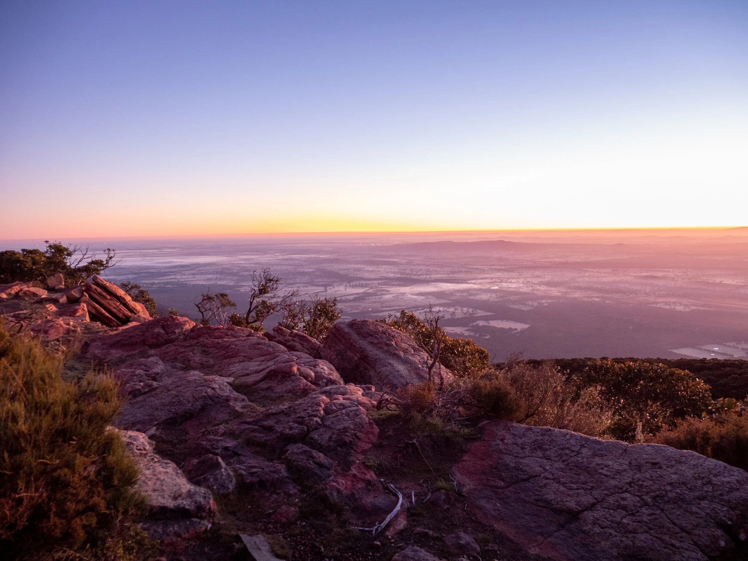 Sunset over a rocky mountain view with trees in the foreground and a flat landscape below.