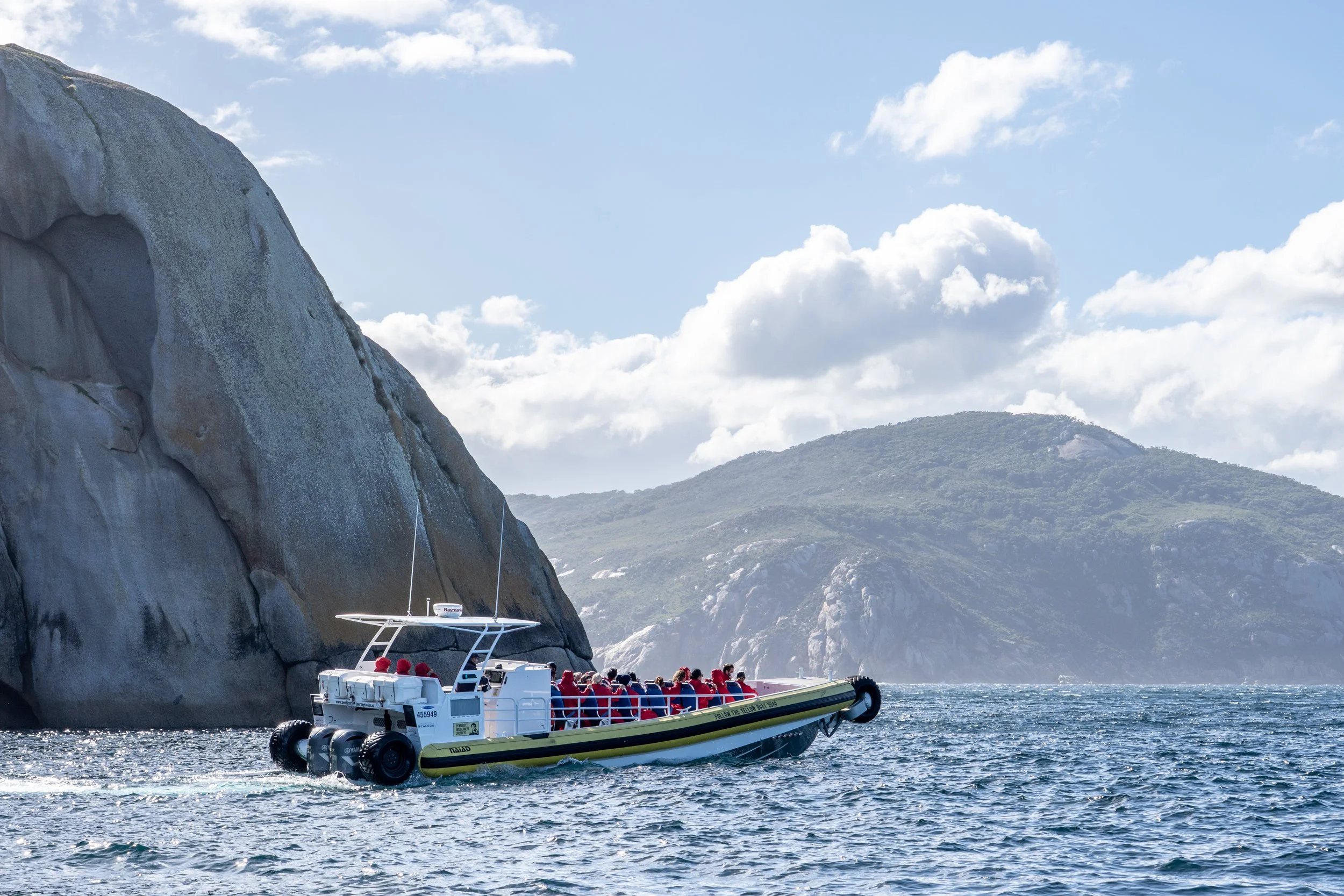 A group of people in red life jackets riding an offshore safety or rescue boat near a large rocky cliff.