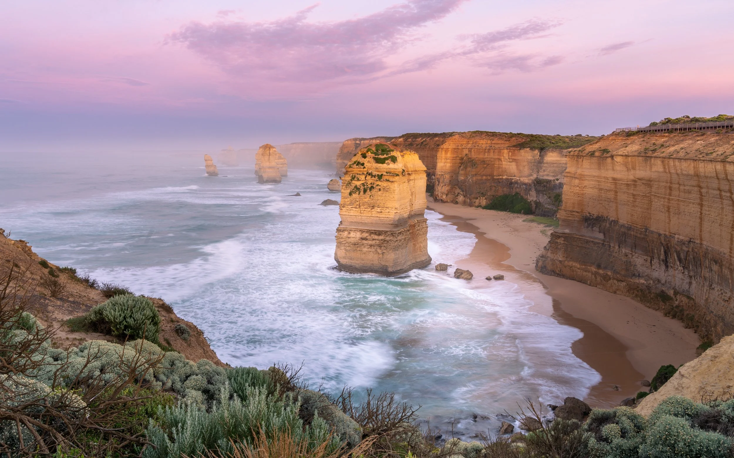 Sunset view of the Twelve Apostles limestone stacks along the coast of Australia with pink sky and waves crashing on the beach.