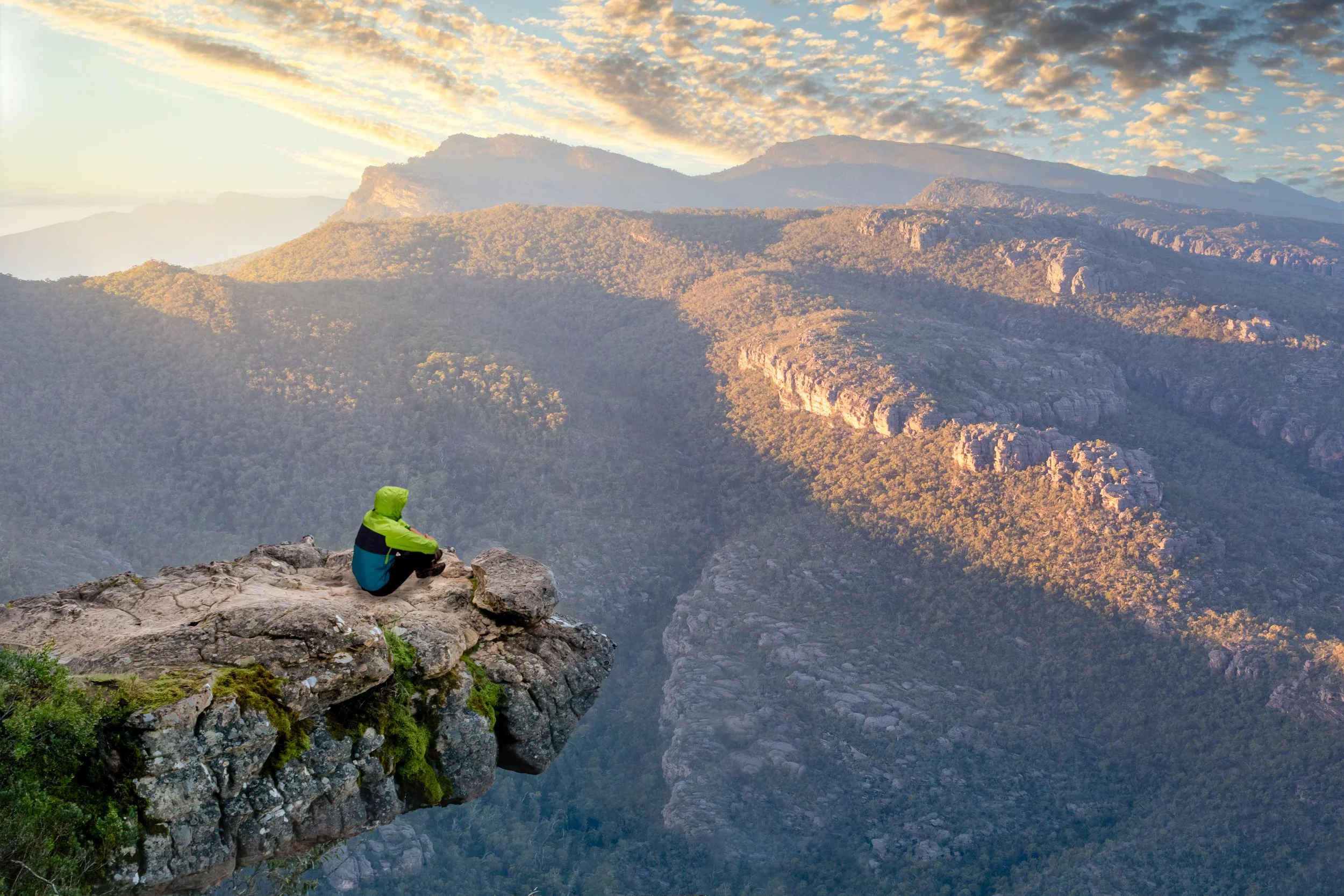 A person in a green jacket sitting on the edge of a rocky cliff overlooking a mountain range during sunrise or sunset.